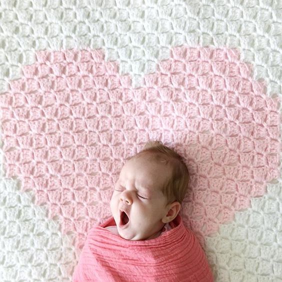 yawning baby swaddled in pink muslin blanket lying on white crochet blanket with pink heart in middle