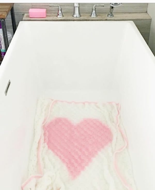 white crochet blanket with pink heart in middle lying in white bathtub