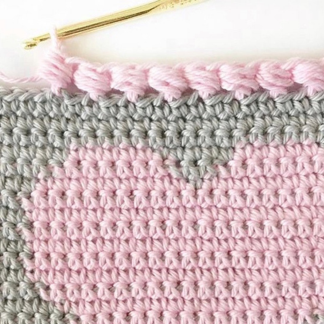 gray crochet blanket with pink heart with pink puff edge around border in progress with gold crochet hook