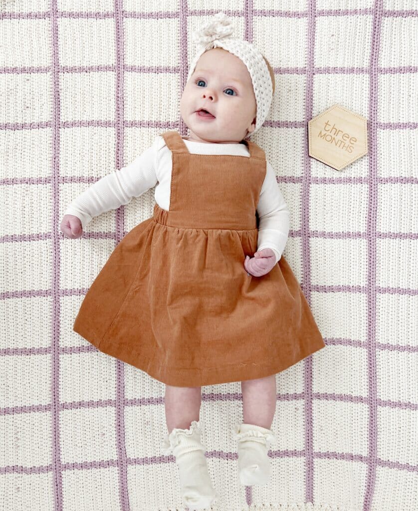Darling baby girl on her 3 month photo shoot laying on a cream and lavender window pane design baby blanket wearing a rust colored dress and a headband. 