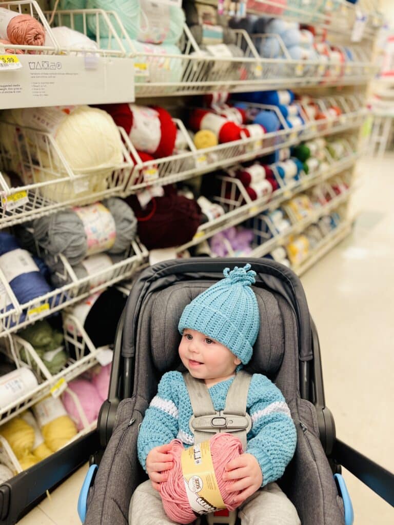 baby in stroller happily smiling at a craft store in the yarn isle. Baby is wearing a crocheted hat and sweater