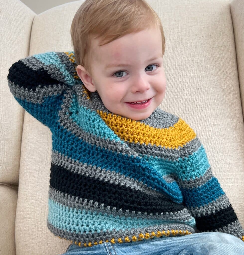 little boy in a crochet striped sweater in teal, grey and black with a pop of gold