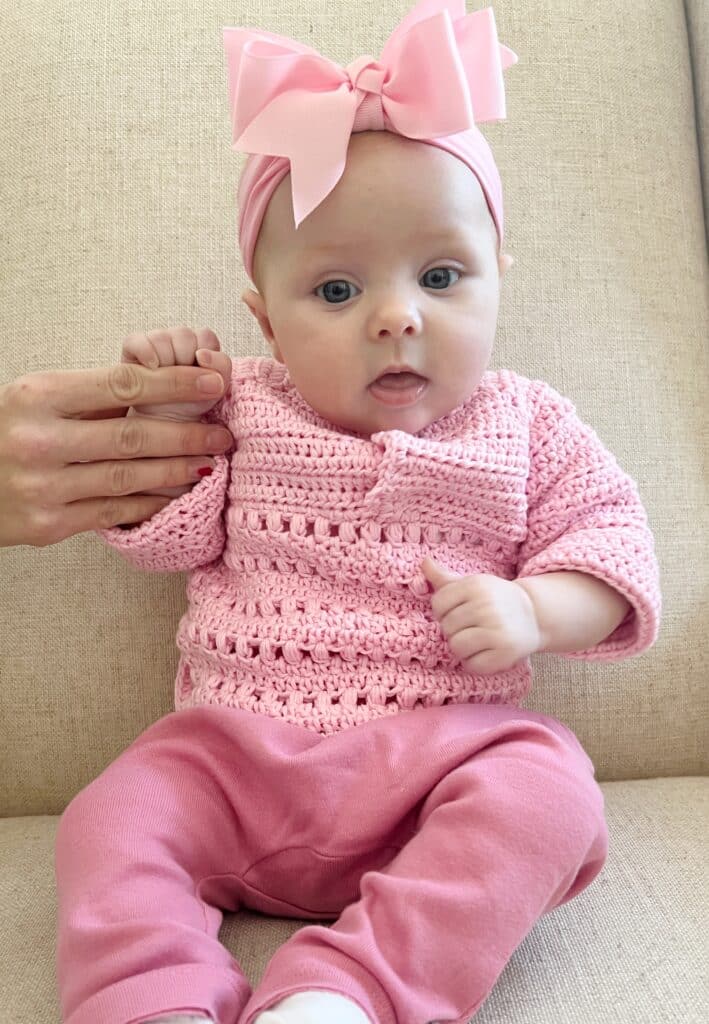baby girl sitting in a cream colored chair wearing a large pink bow, a pink crocheted sweater and a darker pink pair of leggings.