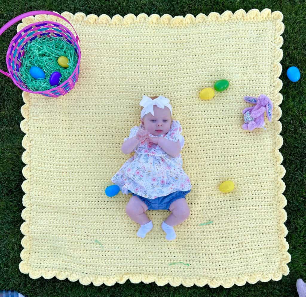 infant laying on yellow crochet blanket with easter basket, easter eggs on the grass