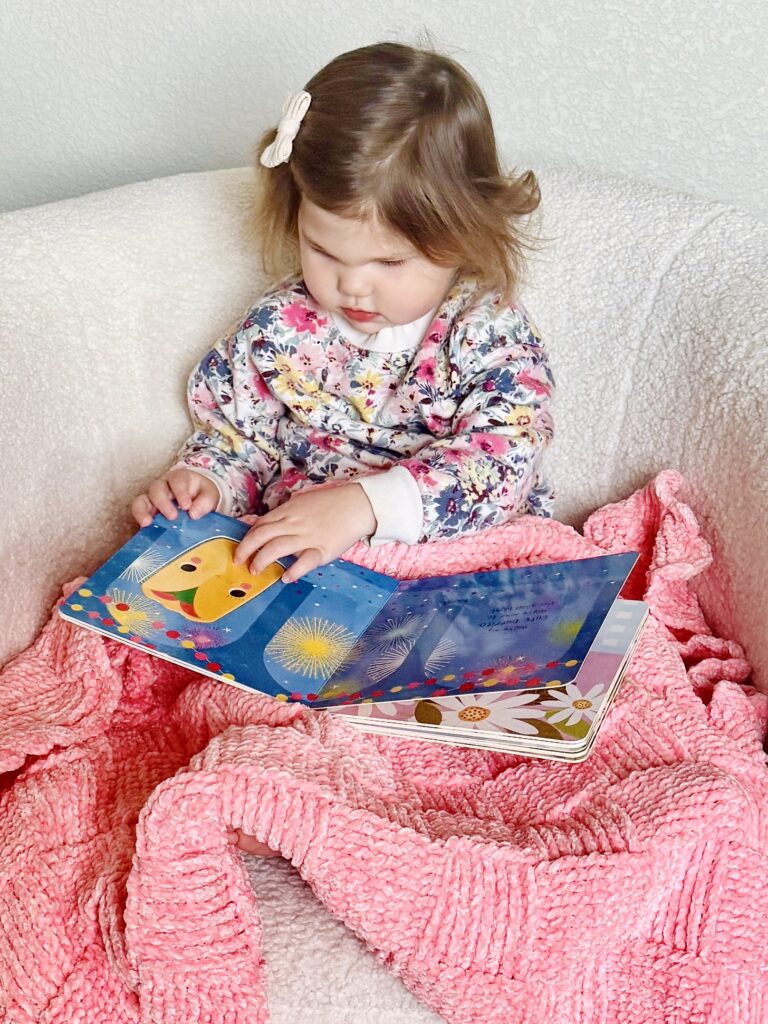 baby girl looking at picture book snuggled under pink crochet blanket